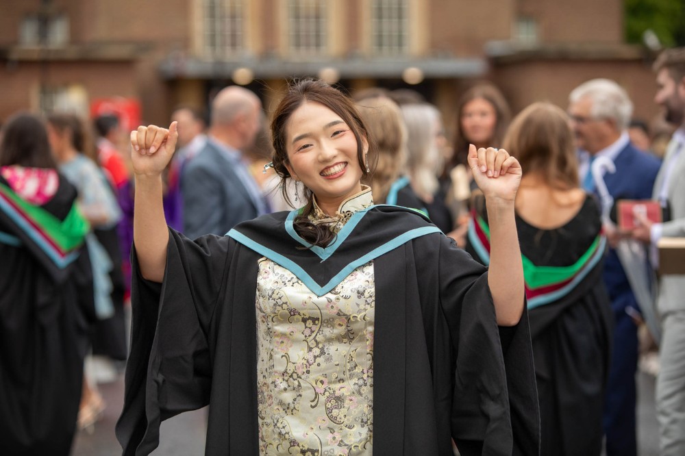 young asian woman in graduation robe making a celebratory gesture with people milling around in the background