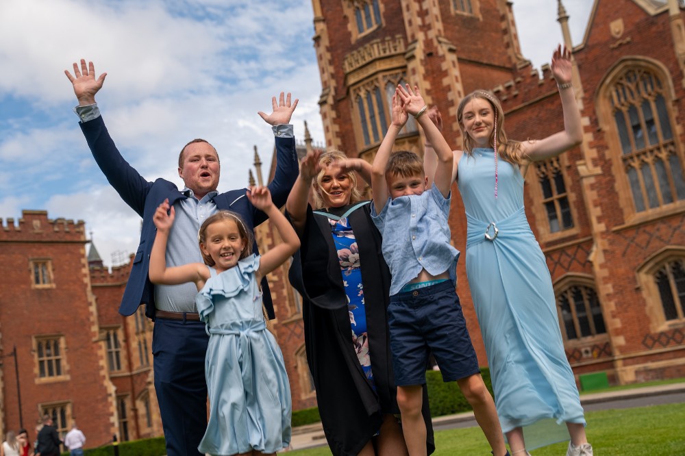 a mum in graduation robes jumping in celebration with her partner and children on a lawn with a redbrick building tower in the background
