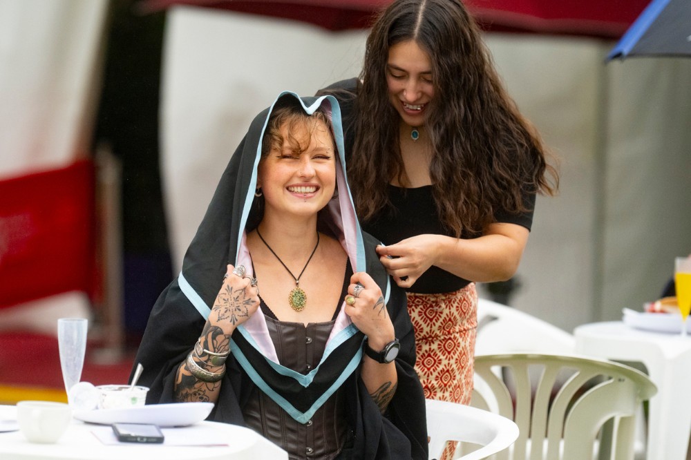 two smiling young women, one seated in graduation garb and the other helping cover her hair to protect it from rain