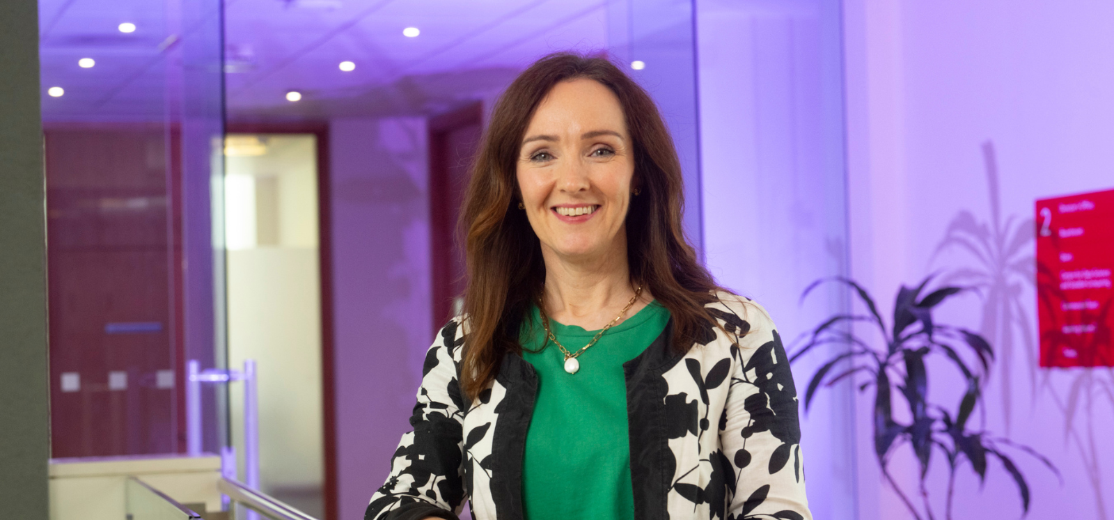 smiling woman wearing green t-shirt and black and white floral-design cardigan in a modern, purple-lit space