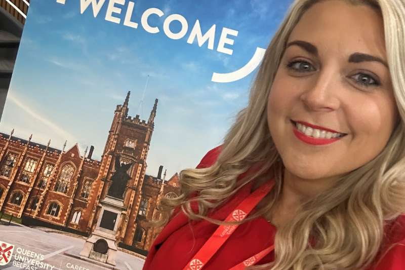 blond-haired woman in red dress smiling in front of a Queen's University Belfast 'Welcome' pop-up stand