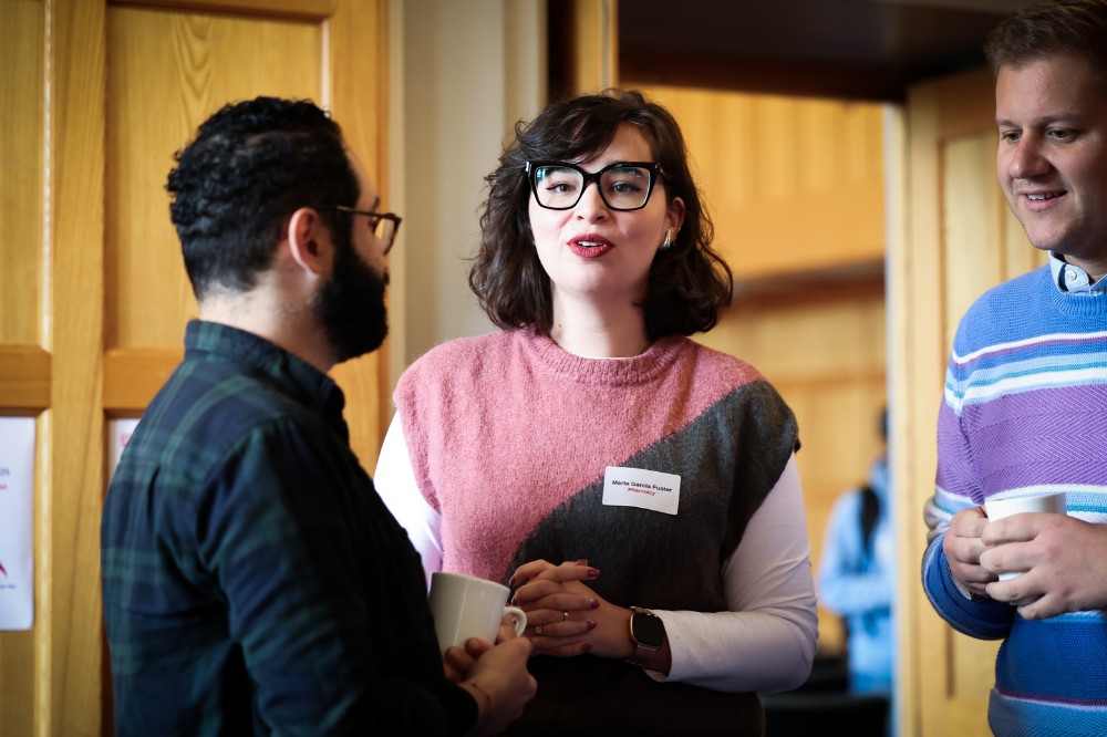 woman with dark-rimmed glasses standing and speaking to two male acquaintances at an indoor event