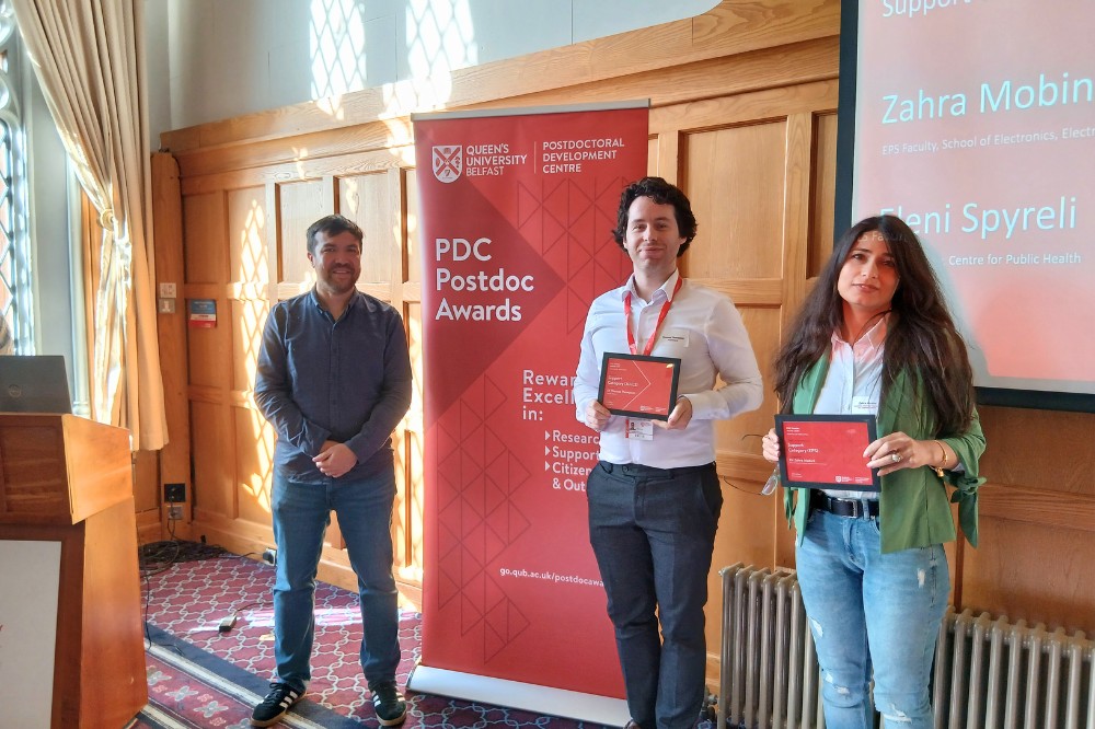three people, two holding award certificates, at an indoor event in a brightly lit, wood-paneled room