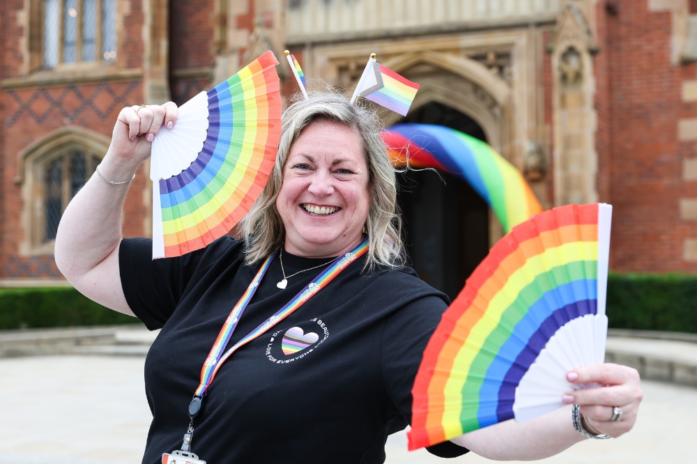 smiling woman in black t-shirt holding two rainbow fans in front of an old redbrick building