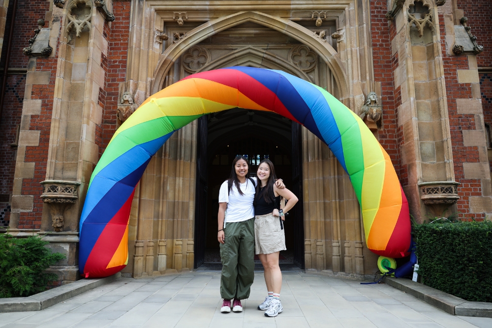 two young women of Asian origin standing under an inflatable rainbow archway at the entrance to an old redbrick building