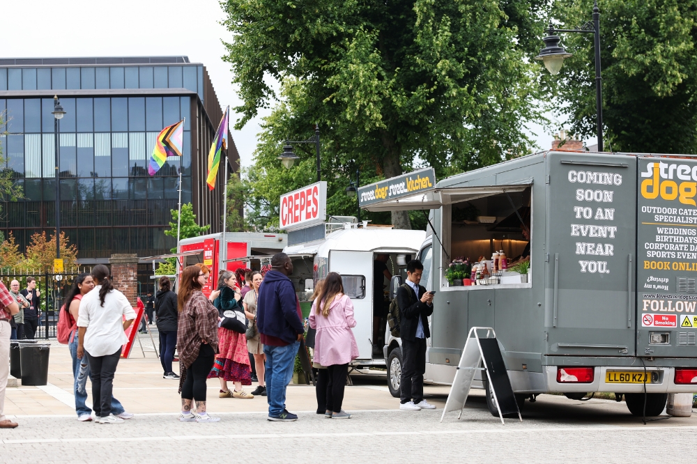 people queueing at a food truck on an open concourse with a modern, glass-fronted building and trees appearing in the background