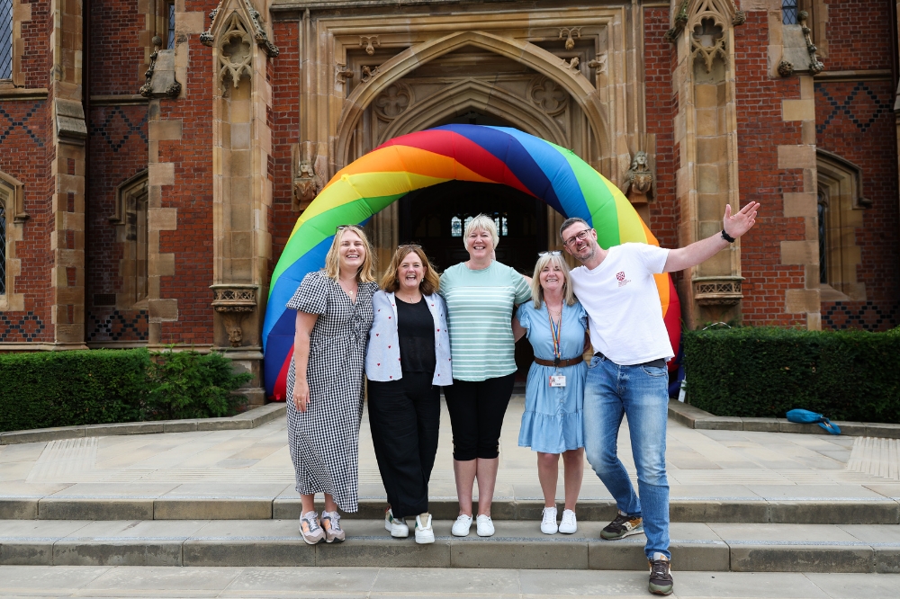 four women and a man smiling to camera in front of an inflatable rainbow archway at the entrance to an old redbrick building