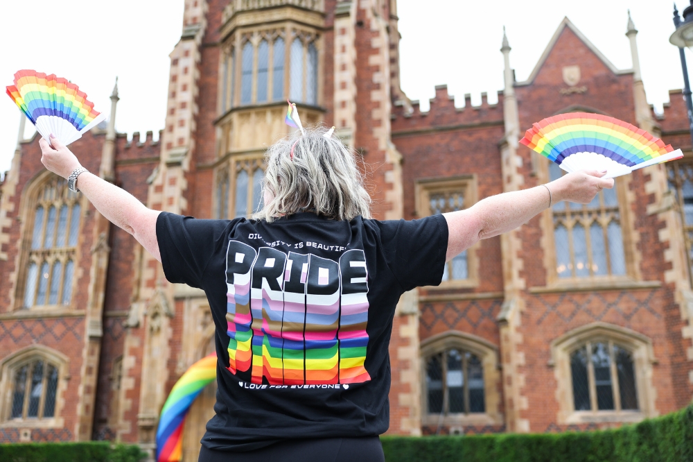 person standing in front of an old redbrick tower wearing a black t-shirt and holding rainbow fans, with their back shown to the camera - t-shirt says PRIDE
