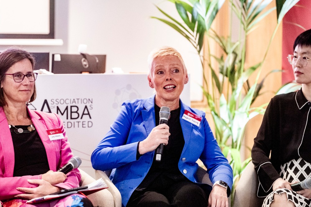 a woman in a bright blue jacket speaking into a microphone as part of a panel, with two other panelists seated either side