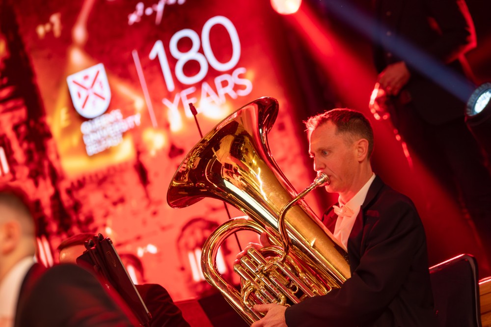 An orchestra tuba player playing at a concert in front of a stage with a presentation screen that reads '180 Years' alongside the Queen's University Belfast logo.