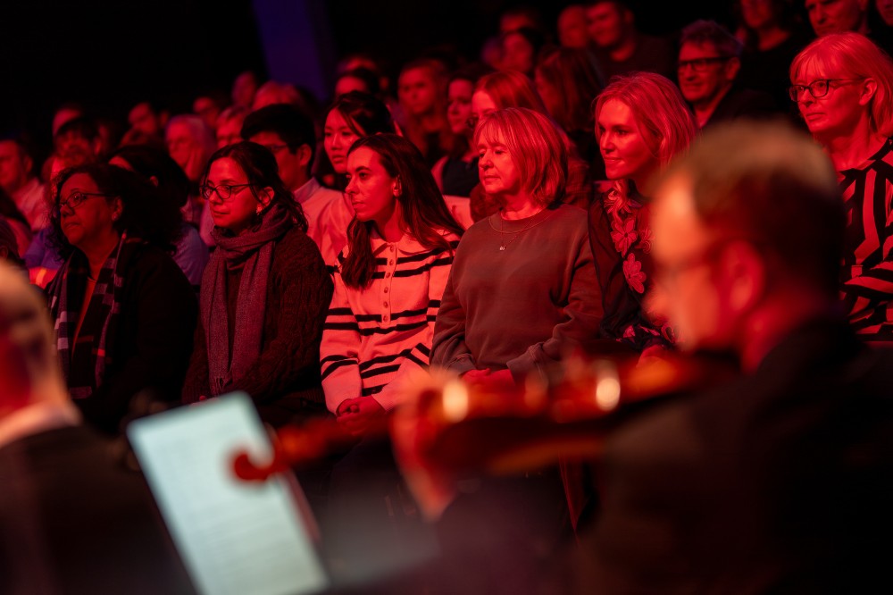 a seated audience pictured listening to a concert, with blurry orchestral players shown in the foreground
