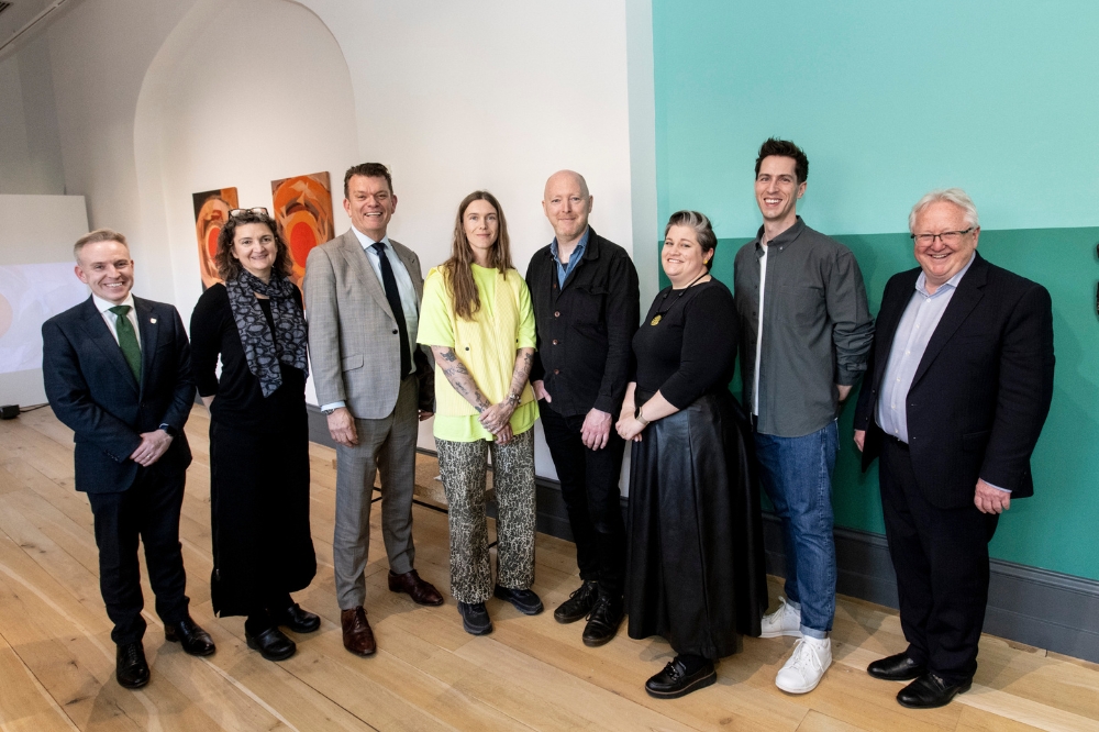Eight members of Queen's staff dressed in smart and casual attire, stand smiling together in a gallery or exhibition space, with artwork displayed on the walls behind them.