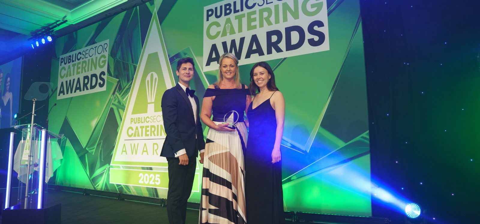 Woman on stage holding an award at a black tie awards ceremony, with man standing on her right and woman standing on her left. Background includes big screen with Public Sector Catering Awards wording and logo, and lectern