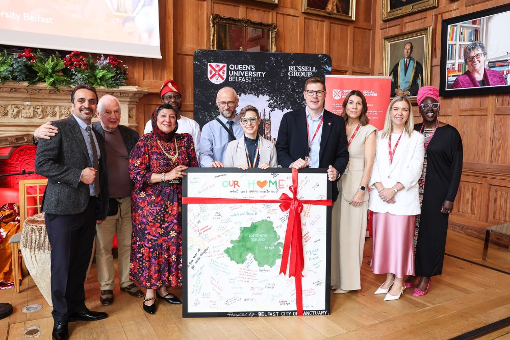 Group of mixed race, gender aged people standing at the front of a large wood paneled room in front of a picture of Northern Ireland featuring signatures and messages.