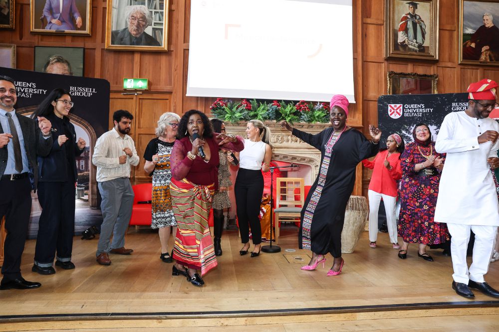 Group of smiling mixed race, age, gender people singing and dancing on a raised dais at the front of a large wood paneled room