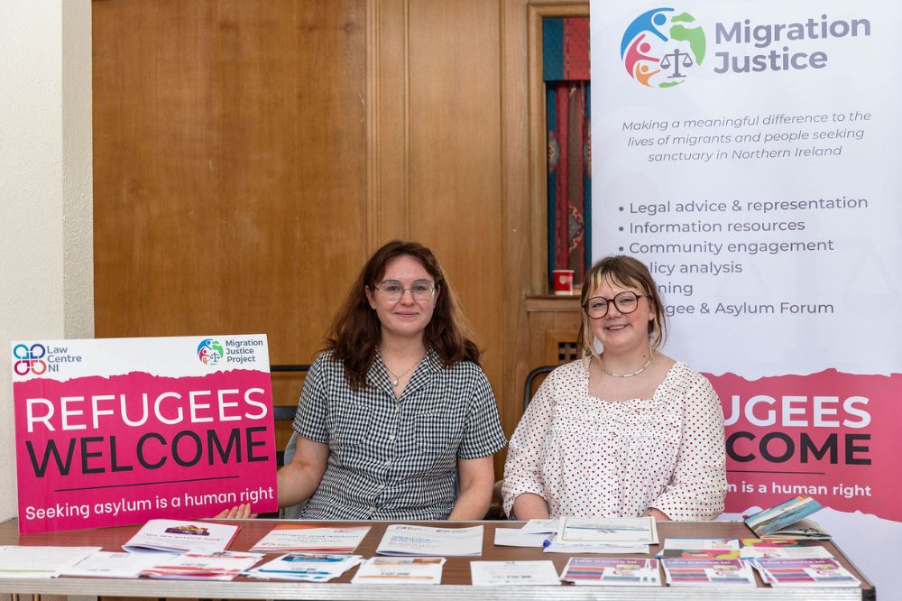 Two ladies sitting in front of an information stand from the Law centre NI on migration justice