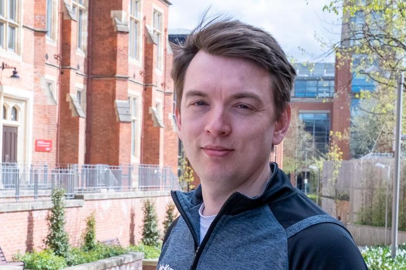 man looking to camera with old redbrick building to the left and modern redbrick building with tower in background, with foliage in shot