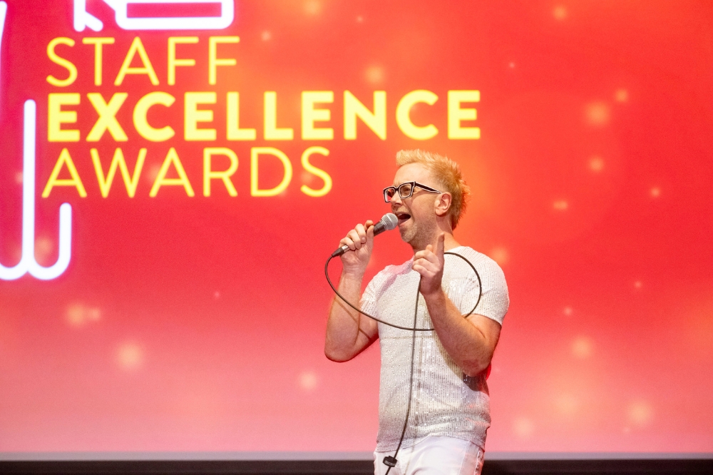 man singing on stage with projection screen in background reading 'Staff Excellence Awards'