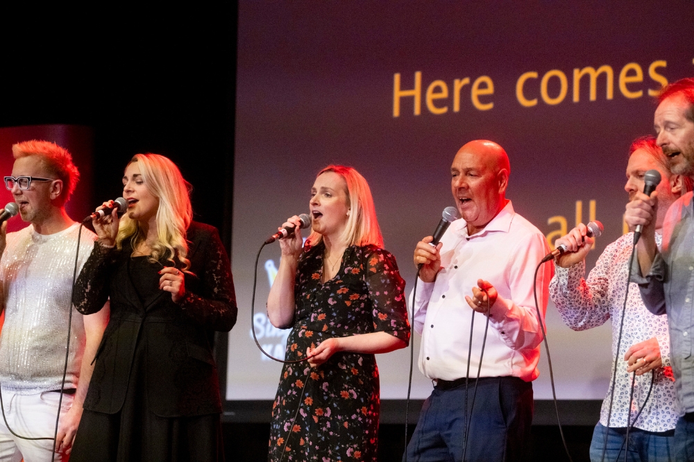 group of people singing on a stage with song words on screen in background