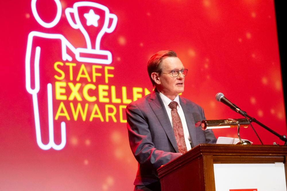 Man in suit speaking from a podium at an event. Background screen reads 'Staff Excellence Awards'.