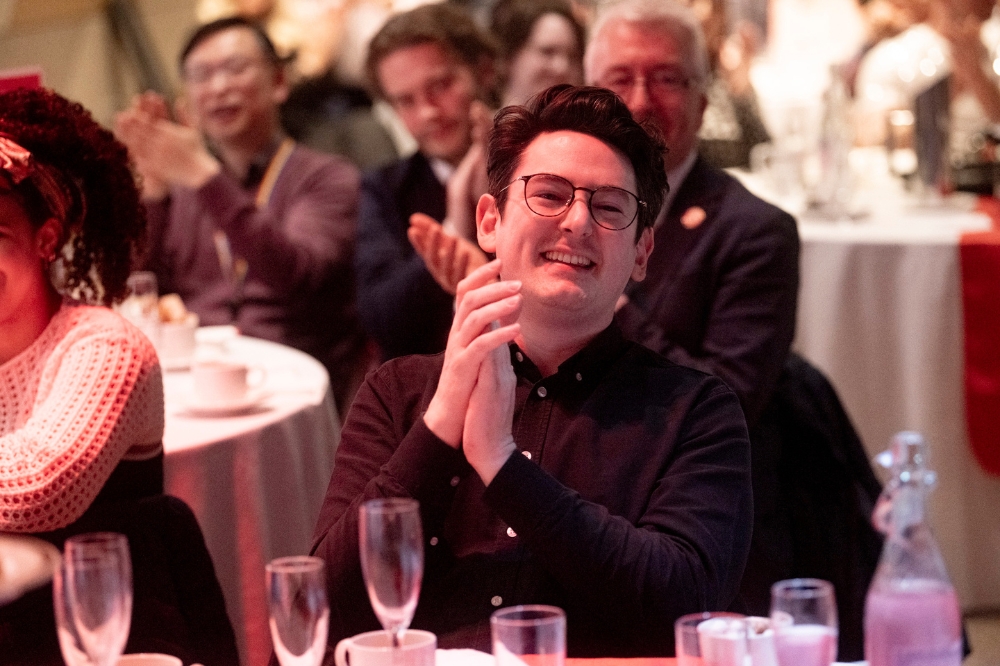 seated bespectacled man applauding and smiling at a dinner table with other event guests in background