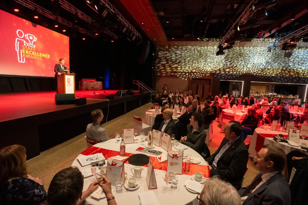 man on stage, left of picture, speaking to a large gathering of people at a gala event