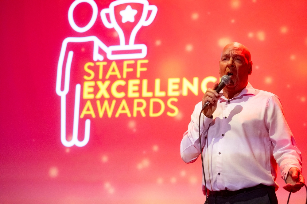 man in white shirt singing on stage. Large background screen reads 'Staff Excellence Awards'.
