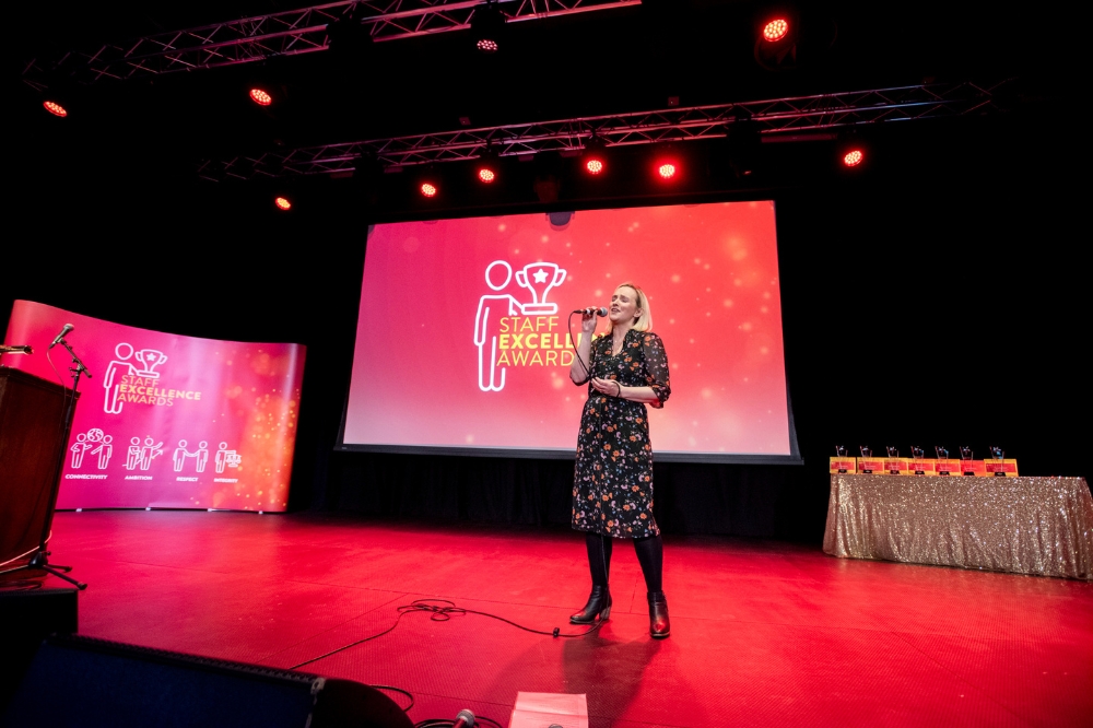 woman singing on stage with large background screens reading 'Staff Excellence Awards'.
