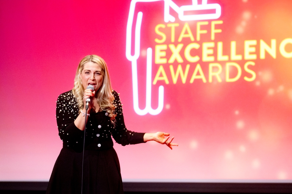woman singing on stage with large background screen reading 'Staff Excellence Awards'.