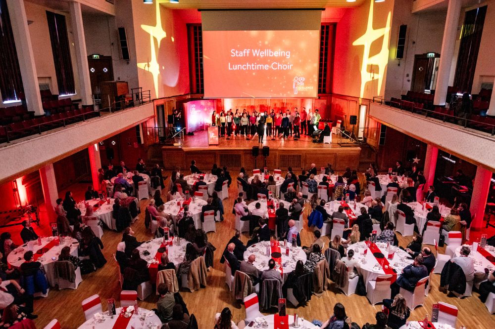 Large hall with groups of people sitting at dining tables listening and watching a performance by a choir. Wording on the presentation screen in the background reads 'Staff Wellbeing Lunchtime Choir'.