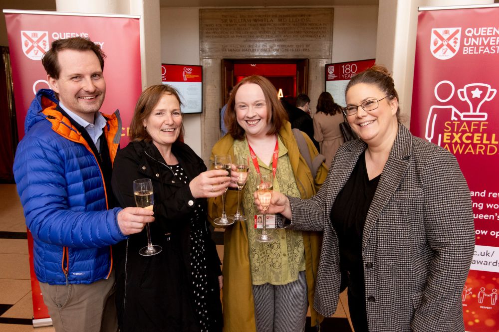Man and three women in celebratory mood posing in the entranceway to an indoor event. A pop-up banner behind the group reads 'Staff Excellence Awards'.