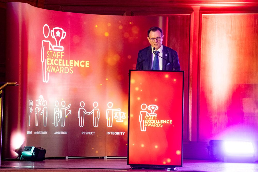 Man in suit speaking from a podium on a stage. The podium façade and a pop-up banner on the stage read 'Staff Excellence Awards'.