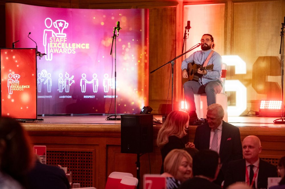 seated guitar player singing and playing on a stage with standing microphones in front of him and a pop-up banner in the left background reading 'Staff Excellence Awards'