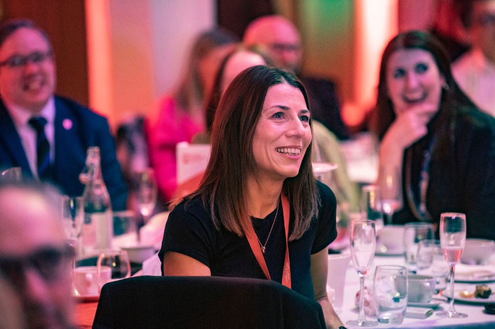 dark-haired woman smiling at a table at an indoor event with other people pictured behind her