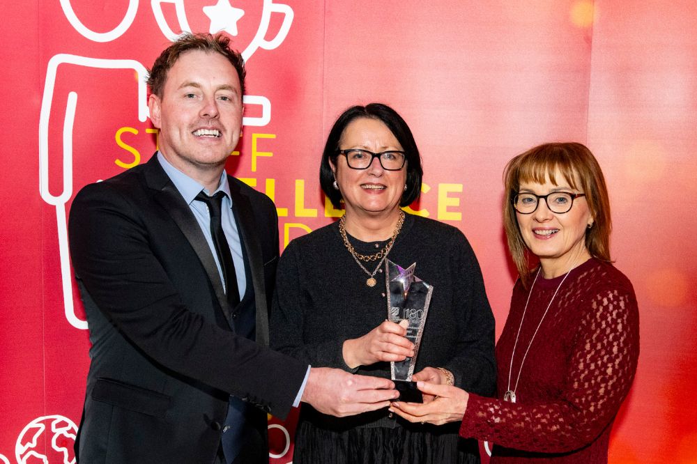 smiling woman in dark dress collecting a crystal trophy award from a man and woman at an event