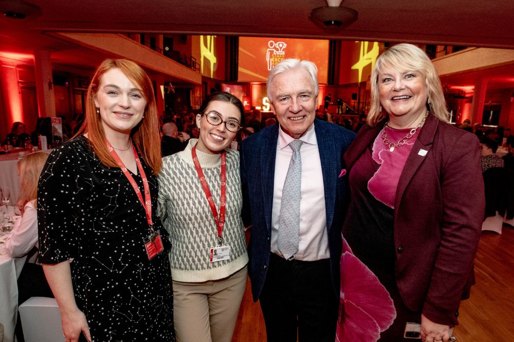man in suit and three casual-smartly dressed women posing for a photograph at an indoor event