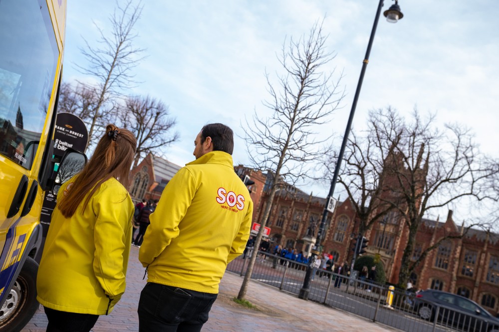 two people in yellow jackets chatting beside a minibus, with an old, Victorian, redbrick building pictured in the background