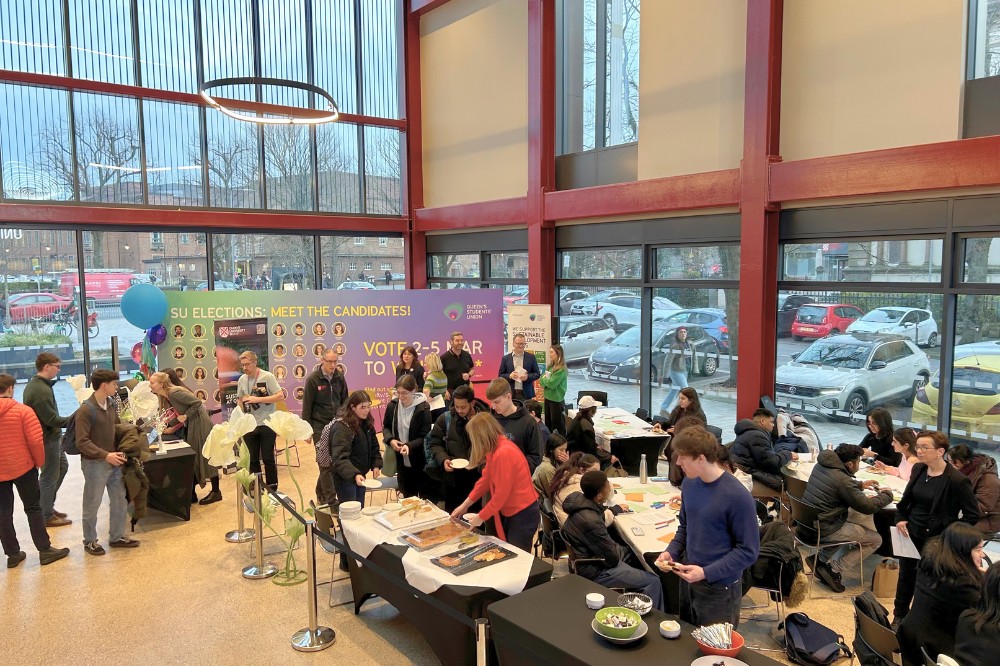 people standing, chatting, taking food from a table, seated and eating at an indoor event with pop-up stands in the background in the atrium of a modern glass-fronted building