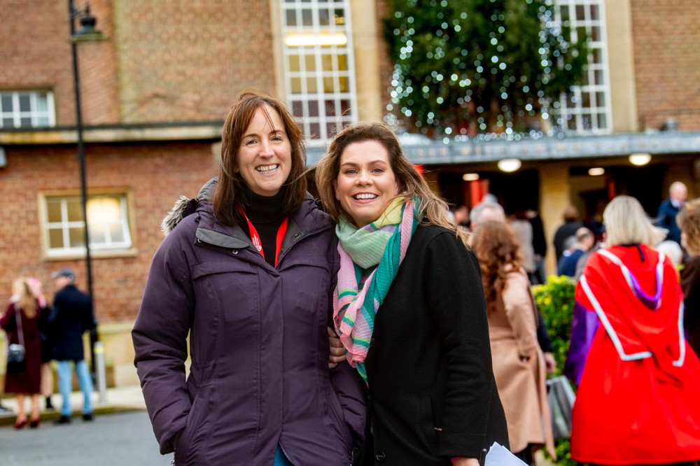 two women in warm clothing smiling to camera with an outdoor Christmas tree pictured in the background