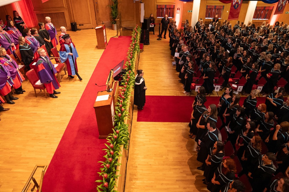 view from the balcony of a graduation ceremony taking place in a hall. Left side of the picture shows the gathered academic procession - right side shows graduands or graduates