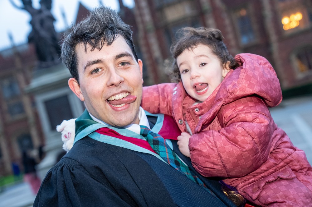 man in graduation robe jokingly sticking his tongue out with his young child in front of an old building
