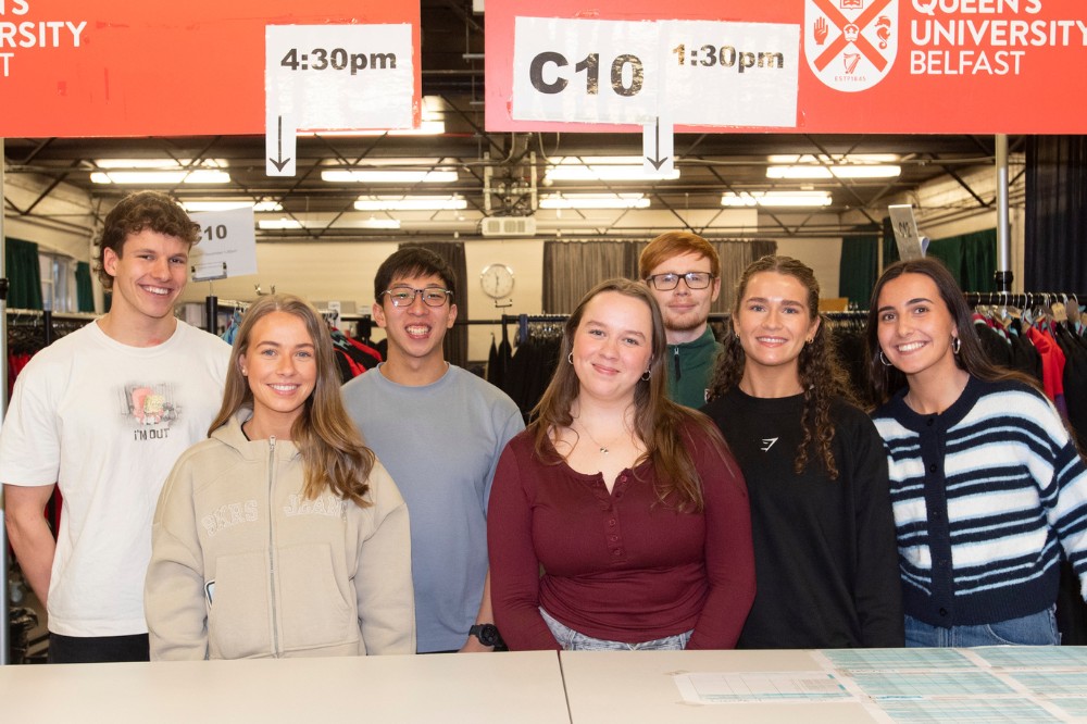 group of smiling young women and men standing at the desk of a graduation gown collection point