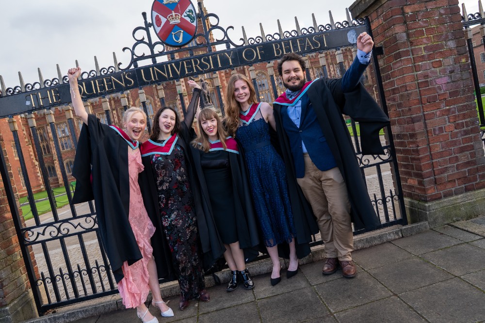 group of four young women and a man celebrating in graduation garb in front of the cast iron gates of Queen's University Belfast